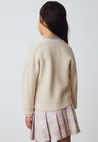 Suéter beige de punto con textura acanalada, cuello redondo y hombros caídos, combinado con una falda plisada de cuadros rosa.