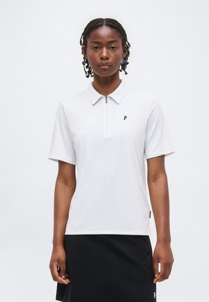 Kvinde med flettet hår iført en hvid kortærmet poloshirt med lynlås og sort nederdel, stående mod en ensfarvet hvid baggrund.