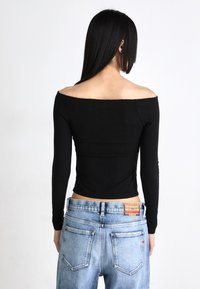 Juodas raukinėtas off-shoulder ilgomis rankovėmis viršus, derinamas su šviesiai mėlynomis džinsinėmis kelnėmis, parodantis trumpą siluetą ir prekės ženklo etikete ant juosmens.