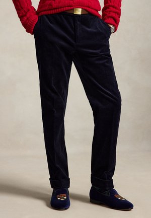 Homme portant un pantalon en velours côtelé noir plié, un pull en maille rouge et des mocassins en velours bleu marine avec un blason brodé, mains dans les poches.