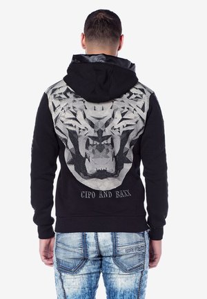 Mann trägt schwarzen Hoodie mit großem geometrischem Tigerkopfdesign und dem Schriftzug "CIPO AND BAXX" auf dem Rücken, kombiniert mit hellblauen Denim-Jeans.