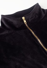 Mustast sametist jakk, millel on osaliselt üles tõmmatud kuldne tõmblukk, näidates kõrget kraed õmmeldud õmbluste detailidega.