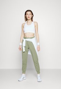Sutiã desportivo branco, calças de chà de cor verde-claro, e uma camisa branca atada. O modelo usa ténis atléticos brancos. Design simples, materiais suaves e corte descontraído.