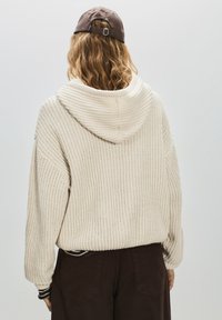 Hanorac tricotat de culoare crem, cu un model texturat, o glugă spațioasă și manșete ribbed. Asortat cu pantaloni maro închis.