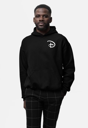 Man met korte baard die een zwarte hoodie draagt met een wit gestileerd "D"-logo en zwarte broek met witte rasterprint.