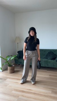 Schwarzer gerippter Rollkragenpullover, graue weit geschnittene Hose, schwarzer Pelzhut, hält eine strukturierte schwarze Tasche, steht in einem gut beleuchteten Raum mit Pflanzen.