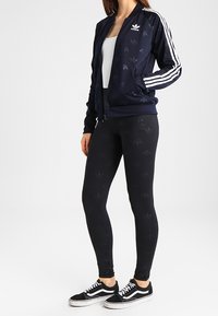 Marinblå träningsoveralljacka med vita ränder, dragkedja fram och subtilt logomönster, tillsammans med svarta leggings med liknande logodesign och svarta sneakers.