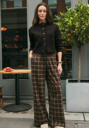 Frau in braunem, geknöpftem Cardigan und weit geschnittenen Karohosen steht mit den Händen in den Taschen vor einem Café neben einem Tisch und einem Topfbaum.