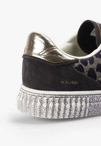 Zapatillas de ante negro con una tapa de talón metálica dorada y suela texturizada gris. Presenta un panel lateral de textil con un patrón y un logo en relieve.