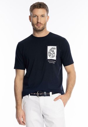 Mężczyzna o krótkich włosach, ubrany w czarną koszulkę z białym wzorem spiralnym i napisem „Marine Club Campione”, białe spodnie oraz pasek.