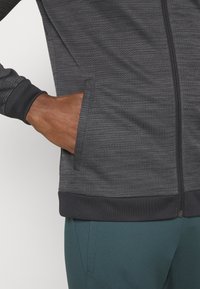 Šedá zipová bunda s texturovanou tkaninou, stojáčkovým límcem, žebrovanými manžetami a boční kapsou s kontrastním černým lemem.