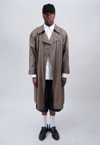 Khaki trench coat, buttoned, with large collar; white shirt underneath; black shorts; white socks; black shoes; plain background.