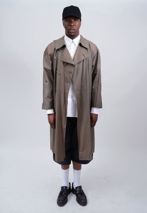 Khaki trench coat, buttoned, with large collar; white shirt underneath; black shorts; white socks; black shoes; plain background.