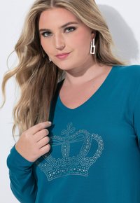 Turkusowa koszula z długim rękawem i dekoltem w serek, z srebrnym wzorem korony wykonanym z cyrkonii na przodzie. Gładka, elastyczna tkanina.