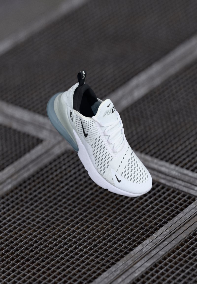 Baskets blanches Nike Air Max 270 avec détails noirs placées sur une surface en grille métallique sombre avec des motifs géométriques.