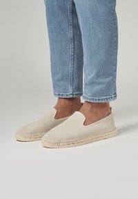 Des chaussures slip-on en suède beige clair avec une semelle en jute tissée. Le design présente des orteils arrondis et un dessus lisse et texturé.