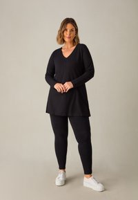 Černý dlouhý rukáv s výstřihem do V a bočními rozparky, v kombinaci s černými legínami a bílými platformovými teniskami na neutrálním pozadí.