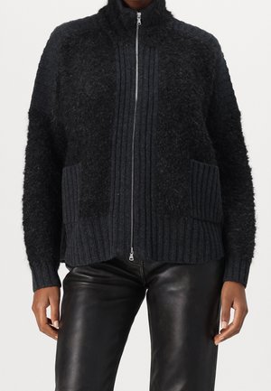 Pull zippé noir texturé avec col, poignets et poches côtelés porté avec un pantalon en cuir noir, montrant le torse et les bras.