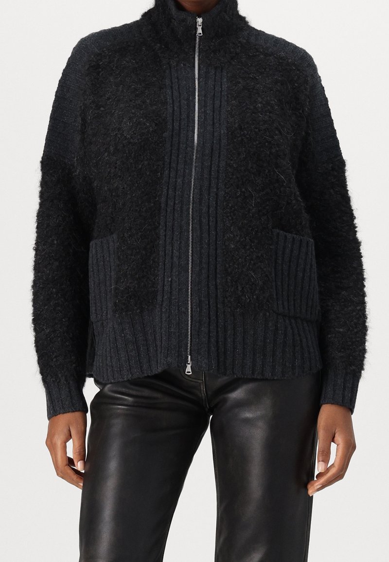 Pull zippé noir texturé avec col, poignets et poches côtelés porté avec un pantalon en cuir noir, montrant le torse et les bras.
