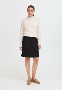 Lichtbeige gebreide trui met een hoge halslijn, gecombineerd met een zwarte jurk tot de knie. Gelaagde stof en eenvoudig ontwerp. Bruine loafers.