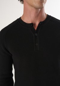 Schwarzes Strick-Henley-Shirt mit langen Ärmeln, das über eine gerippte Textur, eine dreiknöpfige Knopfleiste und ein rundes Kragen-Detail verfügt.