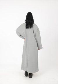 Langer grauer Mantel aus strukturiertem Stoff, mit weiten Ärmeln und geradem Schnitt. Er verfügt über eine Mittelnaht und einen hohen Rücken.