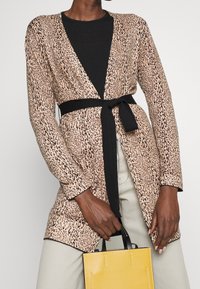 Długi kardigan w panterkę z czarnym paskiem, w połączeniu z czarnym topem, kremowymi spodniami i żółtą torebką. Tkanina o fakturze, otwarty przód.