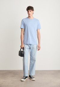 T-shirt azul claro de mangas curtas, ajuste solto, combinado com jeans azul claro. Segura uma pequena mala preta, usando ténis pretos. Fundo simples.