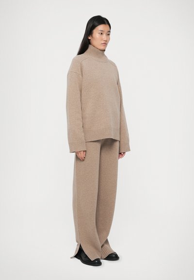 Vrouw die staat en een beige oversized coltrui en wijde gebreide broek draagt met zwarte schoenen tegen een witte achtergrond.