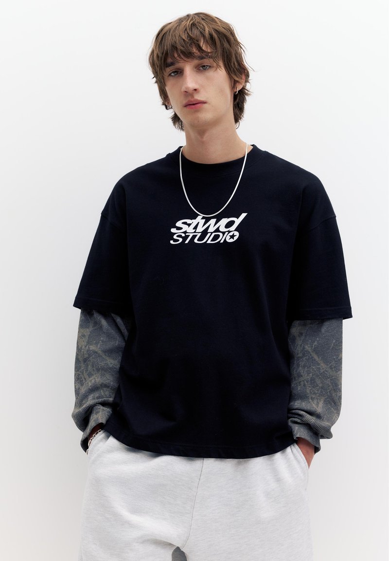 Jeune homme aux cheveux en bataille portant des manches longues superposées noires et grises sous un T-shirt noir à motif graphique et un pantalon gris clair, debout les mains dans les poches.