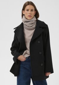 Mustast villast mantel, kahekordse rihmaga, suured kraed ja küljetaskud, kantud helebeige kõrge kaelusega kampsuni ja siniste teksade kohal.