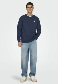 Mørkeblå sweatshirt med rund hals og ribstrikkede manchetter, parret med lyseblå løstsiddende jeans og hvide sneakers. Label synlig på brystet.