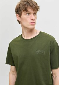 Camiseta de algodón verde oliva con un corte relajado, cuello redondo, mangas cortas y un sutil estampado de logo en la parte frontal del mismo color.