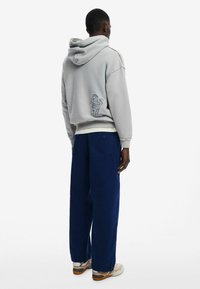 Sudadera gris con puños y dobladillo acanalados, con detalles bordados en la parte posterior. Combinada con pantalones anchos azules y zapatillas beige.