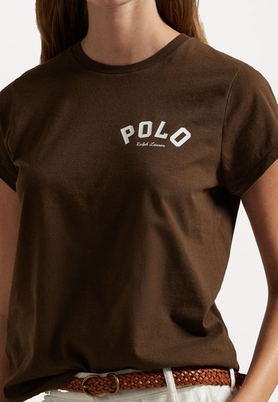 Brązowa koszulka bawełniana z logo "POLO" w białych, łukowatych literach, z krótkim rękawem i okrągłym dekoltem. Noszona z plecionym paskiem.