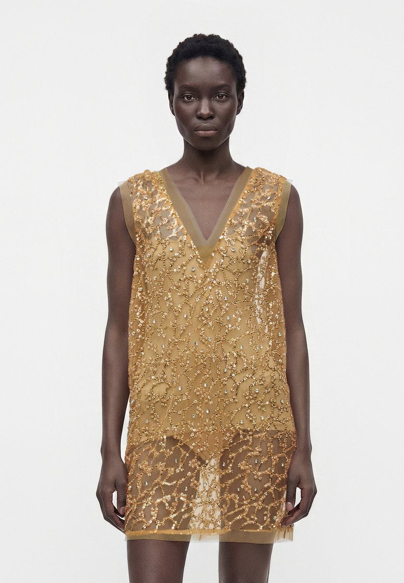 Femme portant une robe dorée sans manches en tissu transparent avec des motifs floraux perlés complexes et un profond décolleté en V, debout devant un fond blanc uni.