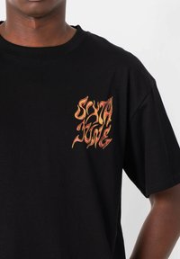 Camiseta negra de algodón con un diseño de texto en llamativo naranja y amarillo "SIXTH JUNE" en la parte superior izquierda del pecho. Mangas cortas, cuello redondo.