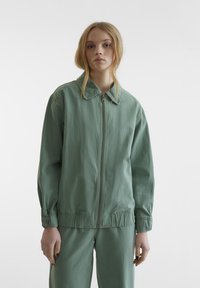 Veste légère verte avec fermeture éclair à l'avant, col pointu et poignets élastiques. Présente une coupe décontractée et une texture subtile.