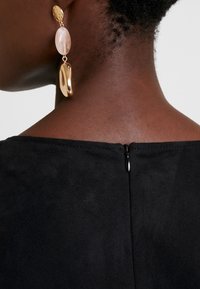 Gros plan sur le cou et l'épaule d'une personne avec une fermeture éclair noire sur le haut et une boucle d'oreille pendante dorée et rose.