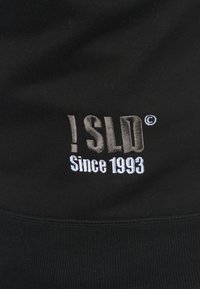 Zwarte sweatshirt met geborduurde tekst: "!SLD ©" in het grijs en "Since 1993" in het wit. Ribbeltextuur langs de boord en de manchetten.