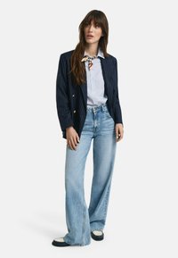 Blazer a righe blu navy con bottoni dorati, camicia azzurra con bottoni, e jeans in denim chiaro a vita alta e gamba larga. Scarpe bianche e blu navy.