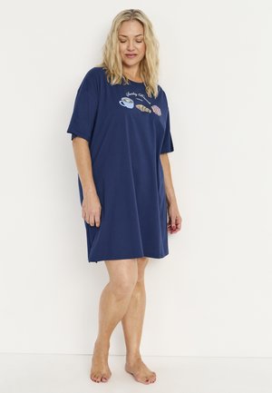 Vrouw met blond haar, gekleed in een losse marineblauwe korte mouw jurk met illustraties van koffie en gebak op de borst, staand op blote voeten.