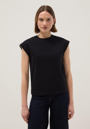 Jonge vrouw met kort bruin haar, gekleed in een zwart mouwloos topje en donkerblauwe jeans, tegen een neutrale achtergrond.