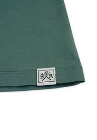 Grøn stofkant med et lille hvidt firkantet mærke med krydsede strikkepinde og bogstaverne B og R.