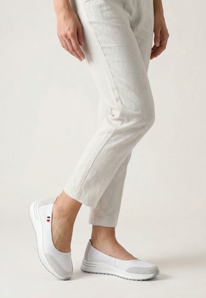 Vrouw draagt lichtbeige cropped broek en witte perforatie slip-on sneakers met een klein vlagdetail aan de zijkant.