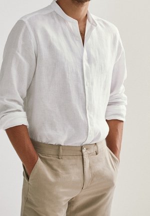 Mężczyzna w białej, zapinanej na guziki koszuli z długim rękawem podwiniętym do łokci oraz w beżowych spodniach, z rękami swobodnie wsuniętymi w kieszenie, na tle gładkiego tła.