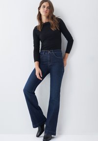 Jeans acampanados de tiro alto en azul oscuro con bolsillos frontales y cierre de botón, combinados con una camiseta negra de manga larga y botas negras hasta el tobillo.