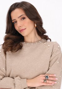 Maglione beige con decorazioni in perle, dotato di scollatura a girocollo e texture lavorata a maglia. Anello dichiarativo in argento e oro su una mano.