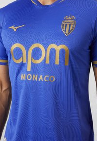Kék sportpóló textúrált körkörös mintákkal, arany logókkal és feliratokkal az "apm" és "MONACO" felirattal, kerek nyakkal, rövid ujjakkal.