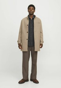 Beige WollmischungsJacke mit Kragen, Knopfverschluss, seitlichen Taschen, kombiniert mit einem dunklen Hemd und braunen Hosen, frontal präsentiert.
